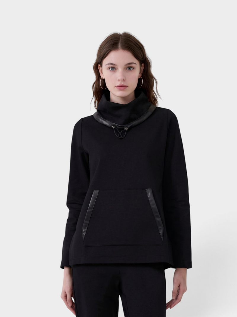 A model wearing a black cowl-neck pullover sweater with glossy black trim details on the collar and pocket edges, paired with black trousers, against a light gray studio background.